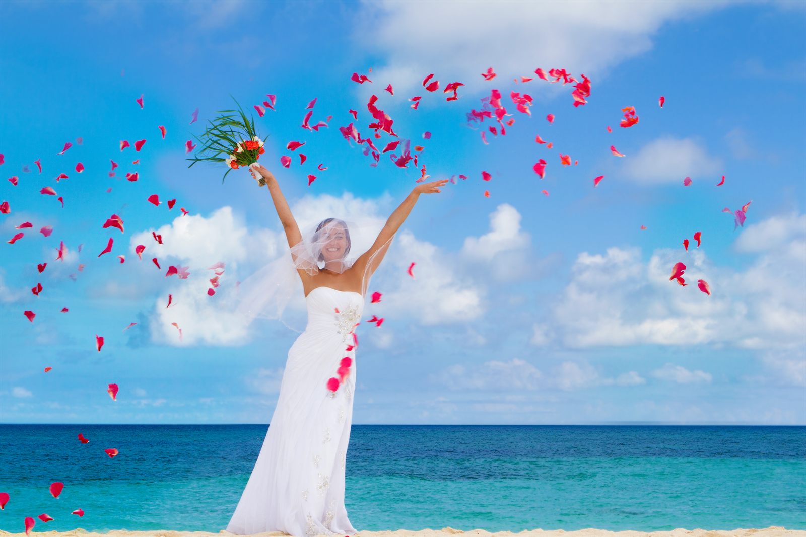 Bride in a white dress throwing red rose petals into the air on a sunny beach
